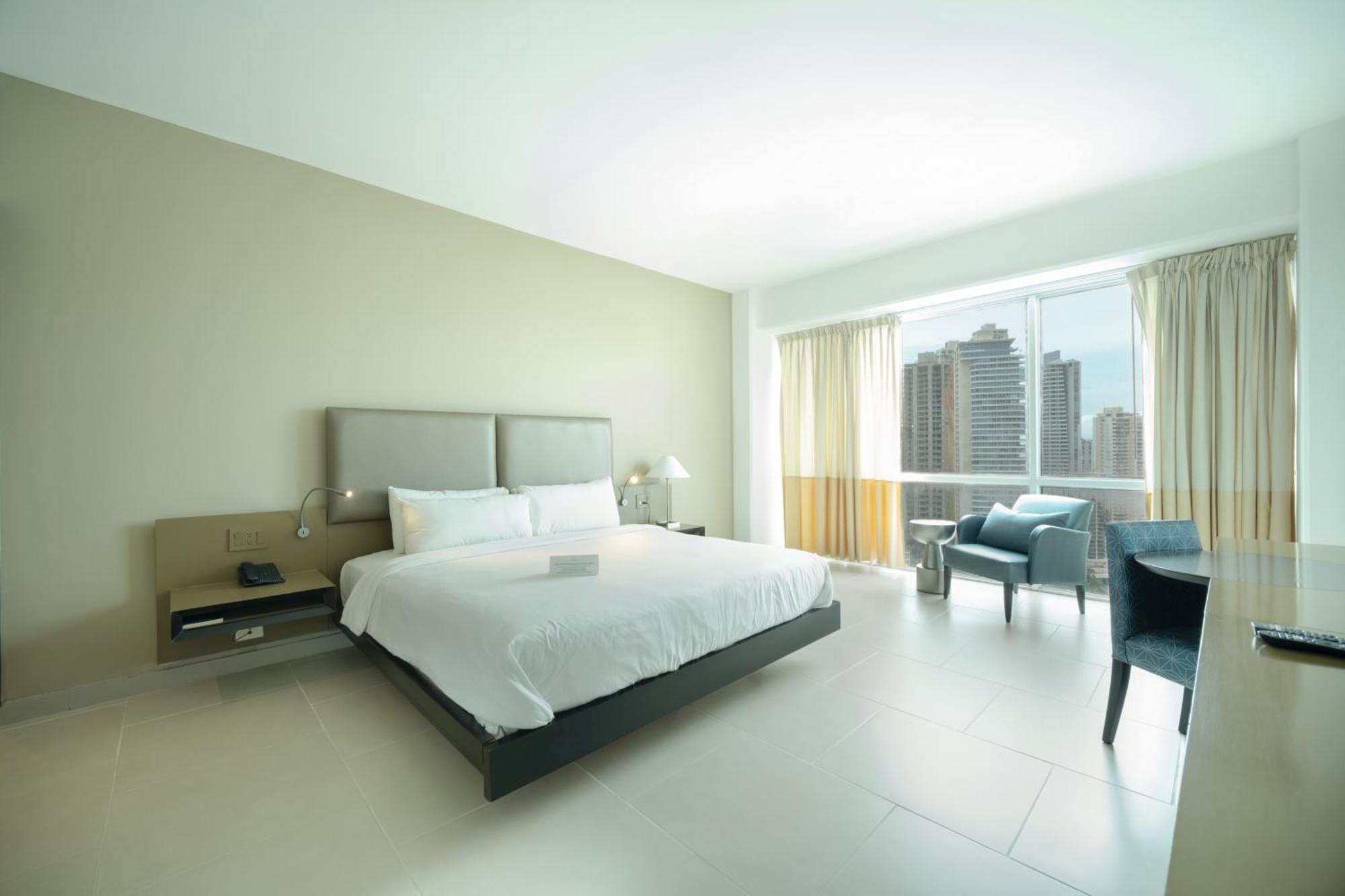Decapolis Hotel ปานามาซิตี้ ภายนอก รูปภาพ The photo shows a modern hotel room with a minimalist design. There is a large bed with a light-colored duvet and two decorative pillows. Next to the bed, there is a nightstand with a lamp. On the opposite side, a comfortable chair is placed near a l