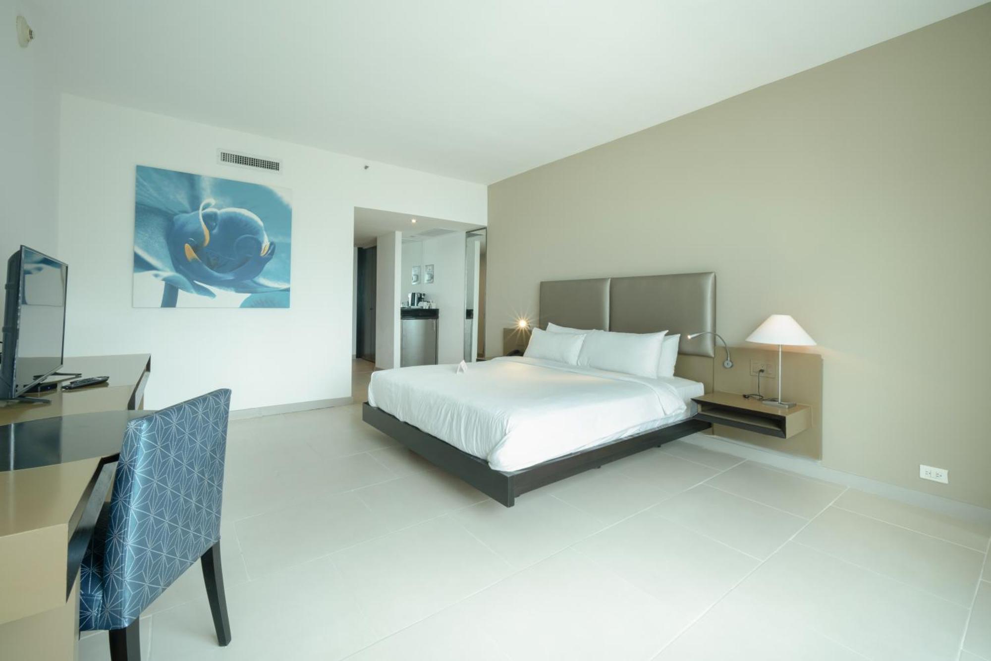 Decapolis Hotel ปานามาซิตี้ ภายนอก รูปภาพ The photo shows a modern hotel room featuring a large bed with white linens. There is a minimalist design with light-colored walls, and a piece of artwork depicting a blue fish on one wall. To the side, there’s a small bedside table with a lamp, and 