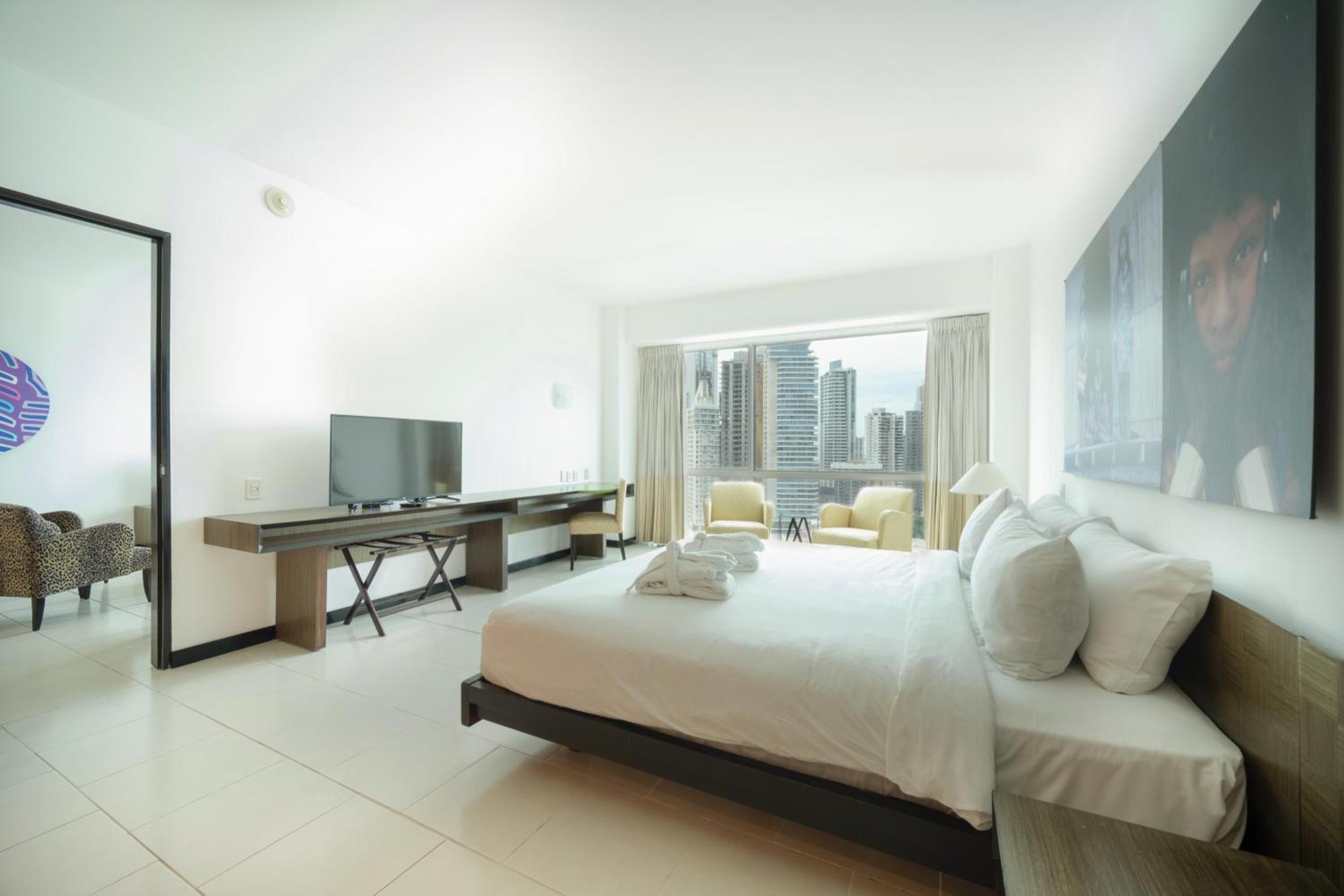 Decapolis Hotel ปานามาซิตี้ ภายนอก รูปภาพ The photo depicts a modern hotel room featuring a large bed with white linens and several pillows. There is a television mounted on the wall opposite the bed, and a desk with a chair beside it. Large windows provide a view of a city skyline, with two