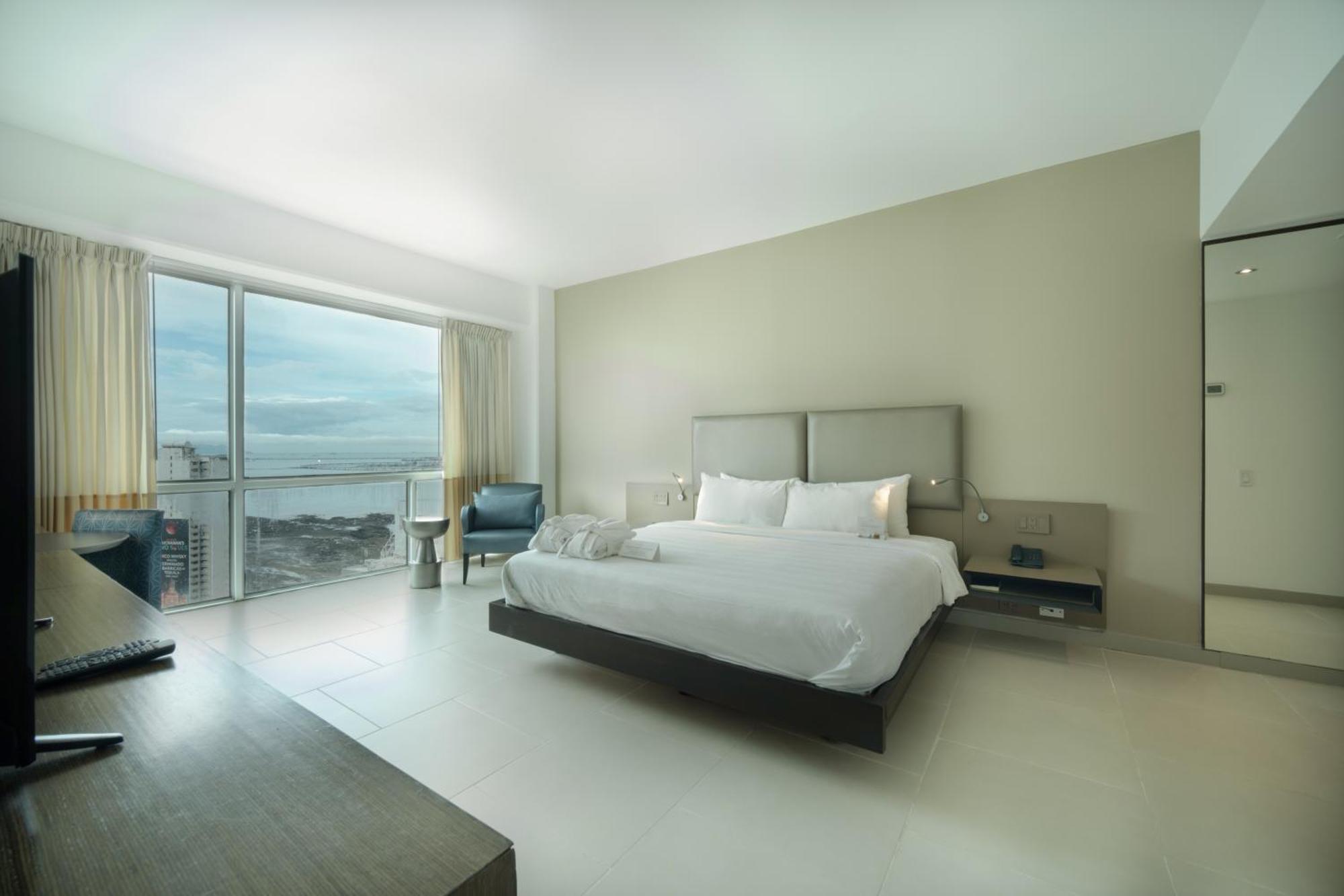 Decapolis Hotel ปานามาซิตี้ ภายนอก รูปภาพ The photo shows a modern and minimalist hotel room. It features a large bed with white bedding, complemented by neutral-colored walls. There is a sleek wooden desk and a chair in the corner, along with a small seating area by a window that looks out 