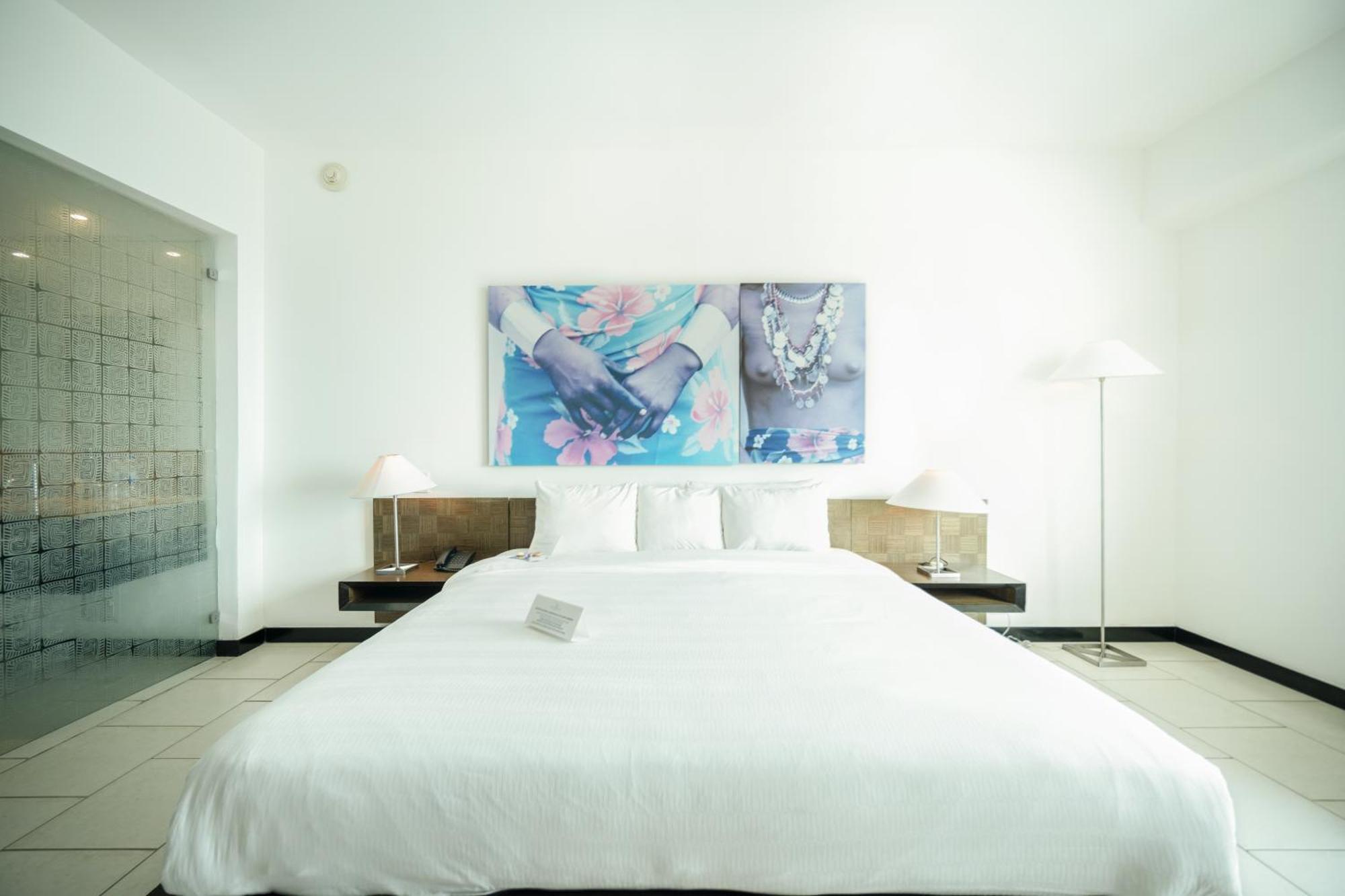 Decapolis Hotel ปานามาซิตี้ ภายนอก รูปภาพ The photo shows a modern bedroom with a clean and minimalist design. The bed is large with a white comforter and several pillows. On the wall above the bed, there are two framed art pieces featuring floral and jewelry motifs, adding a touch of color 