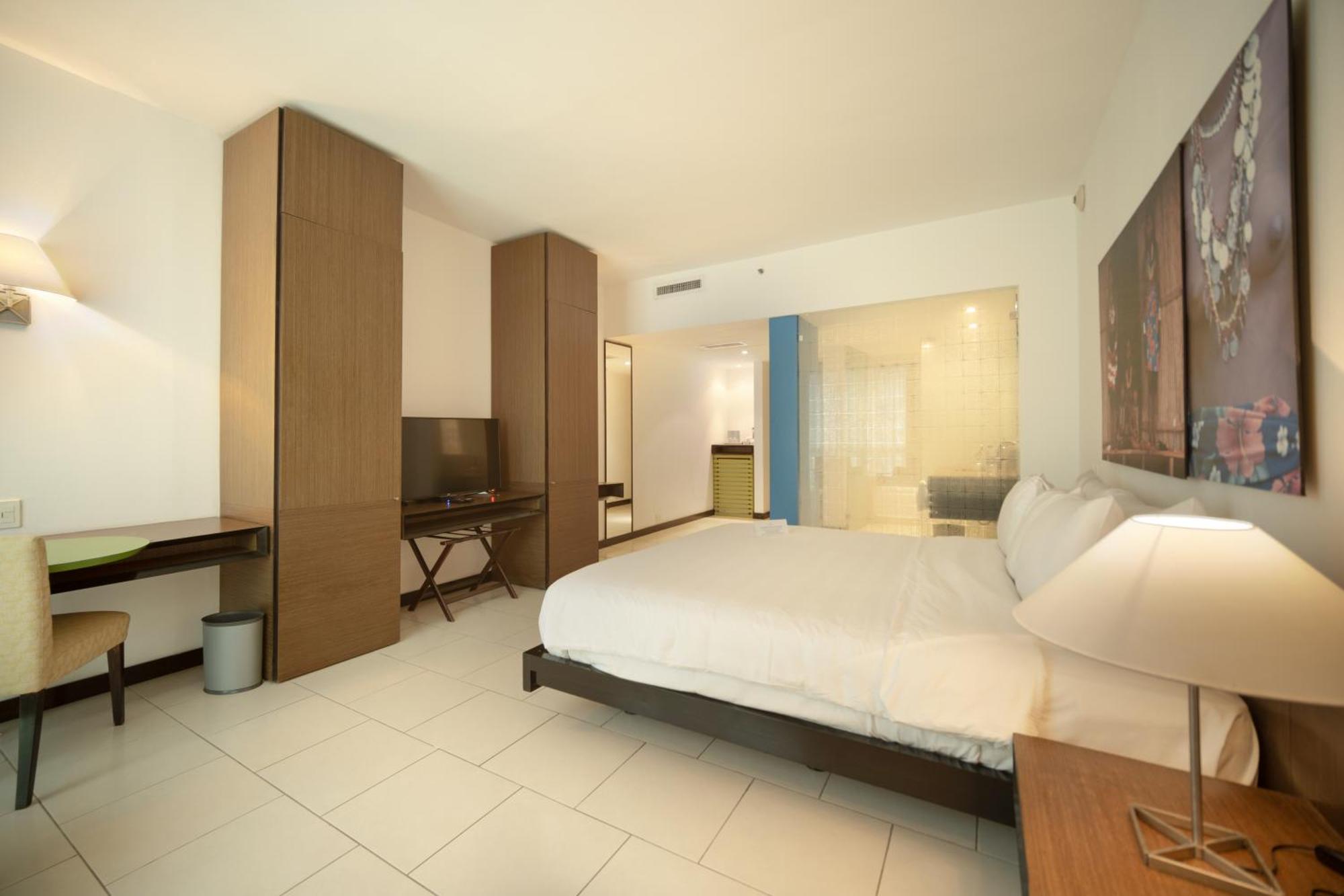 Decapolis Hotel ปานามาซิตี้ ภายนอก รูปภาพ The photo shows a modern hotel room. It features a large bed with white bedding, a wooden headboard, and bedside tables with lamps. There is a wooden wardrobe or storage unit on one side, along with a desk and a chair. A flat-screen TV is mounted on 