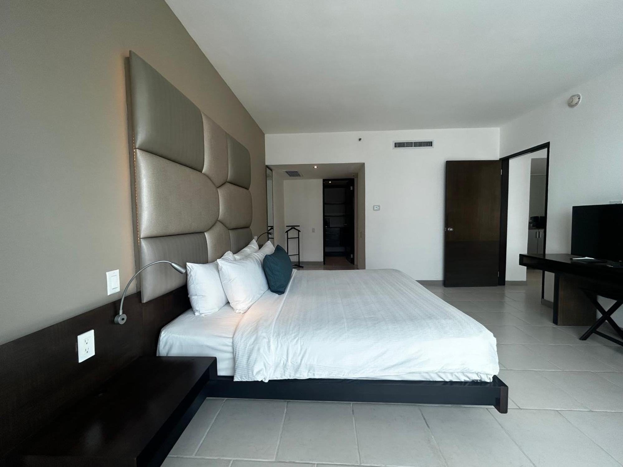 Decapolis Hotel ปานามาซิตี้ ภายนอก รูปภาพ The photo shows a modern hotel room. It features a large bed with a white comforter and several pillows, including a couple of blue accent pillows. The headboard of the bed is upholstered in a light gray material. Next to the bed, there is a dark woo