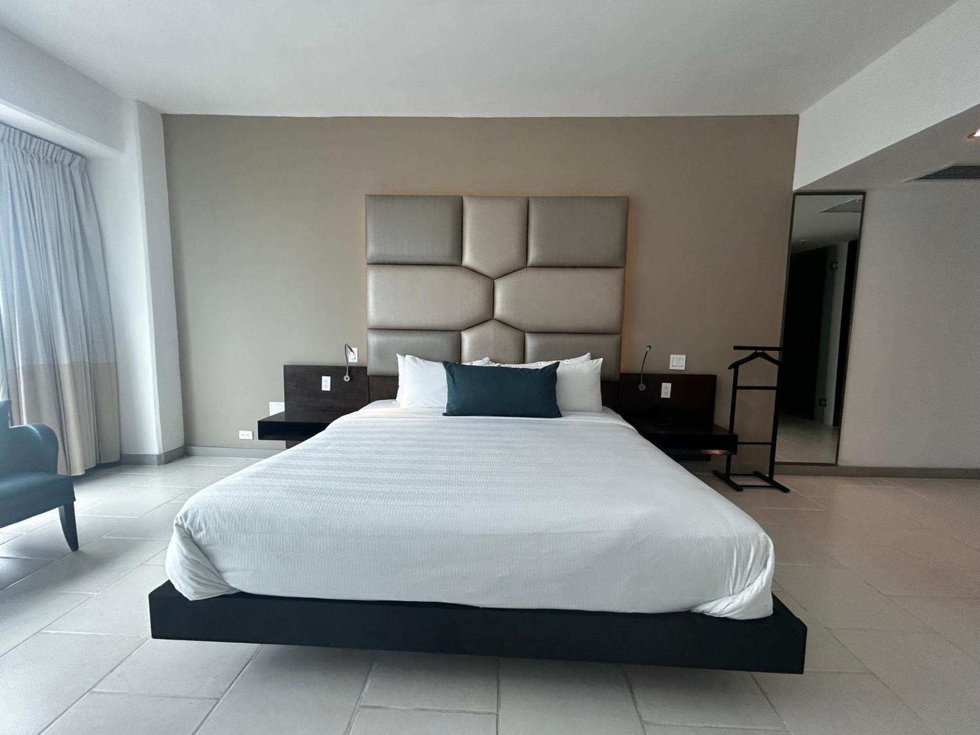 Decapolis Hotel ปานามาซิตี้ ภายนอก รูปภาพ The photo shows a modern and minimalist hotel room. In the center, there is a large bed with a simple white bedspread and a dark accent pillow. The headboard is composed of light-colored padded panels. On either side of the bed, there are dark wooden