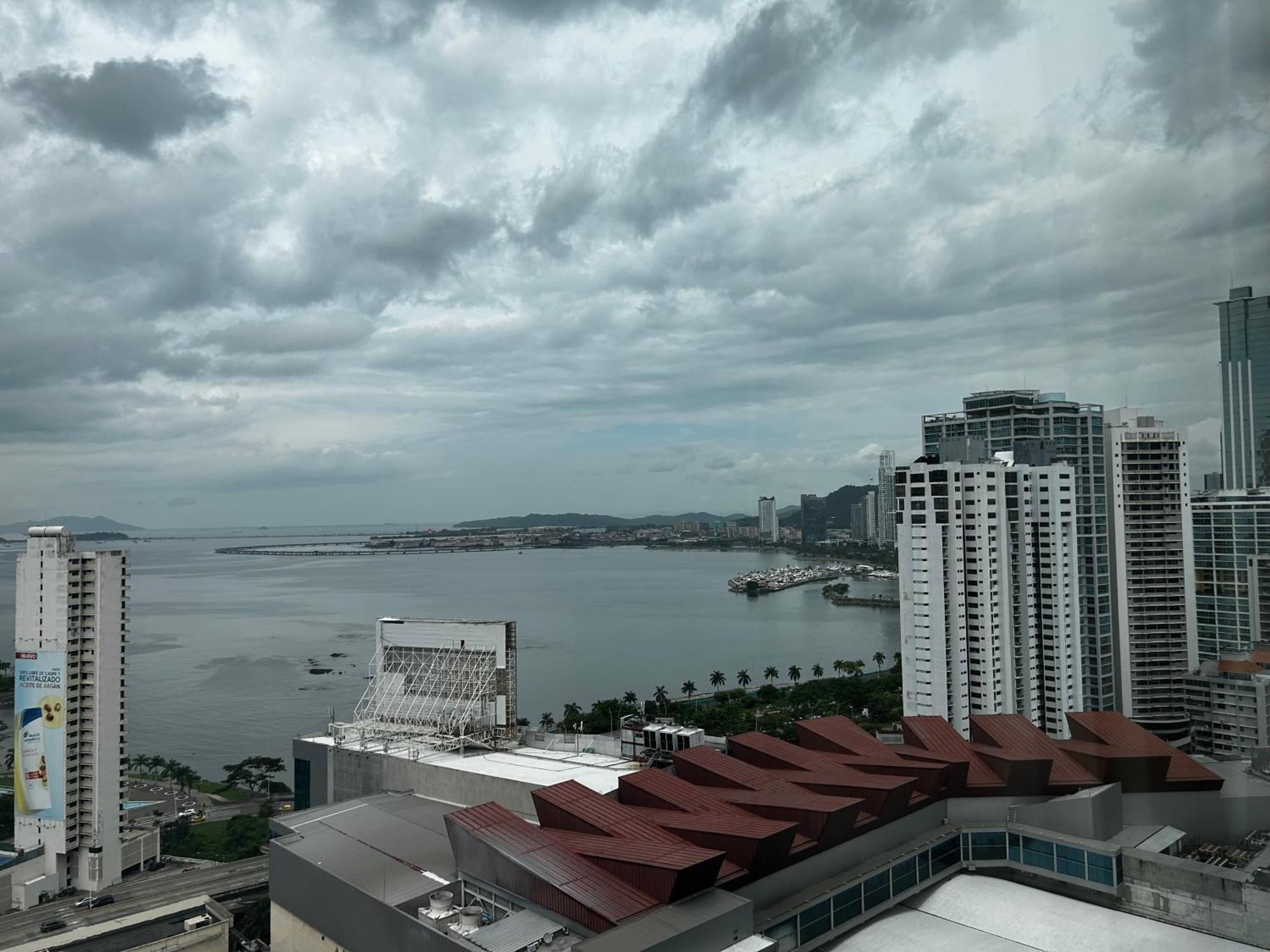 Decapolis Hotel ปานามาซิตี้ ภายนอก รูปภาพ The photo depicts a coastal cityscape under a cloudy sky. In the foreground, there are several modern buildings, including some with distinctive architectural features, like a red-tiled roof. In the background, a bay or harbor can be seen, with water