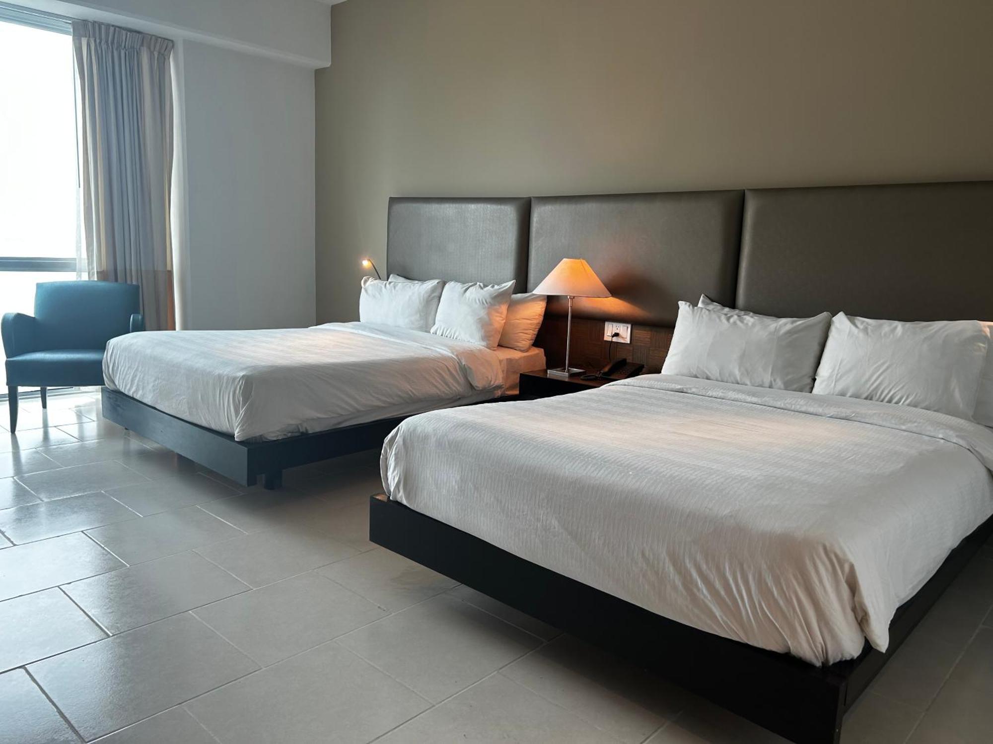 Decapolis Hotel ปานามาซิตี้ ภายนอก รูปภาพ The photo shows a hotel room featuring two large beds with white beddings. There is a bedside table with a lamp next to one of the beds. A cozy chair is placed in the corner of the room. Large windows allow natural light to fill the space, and the wa