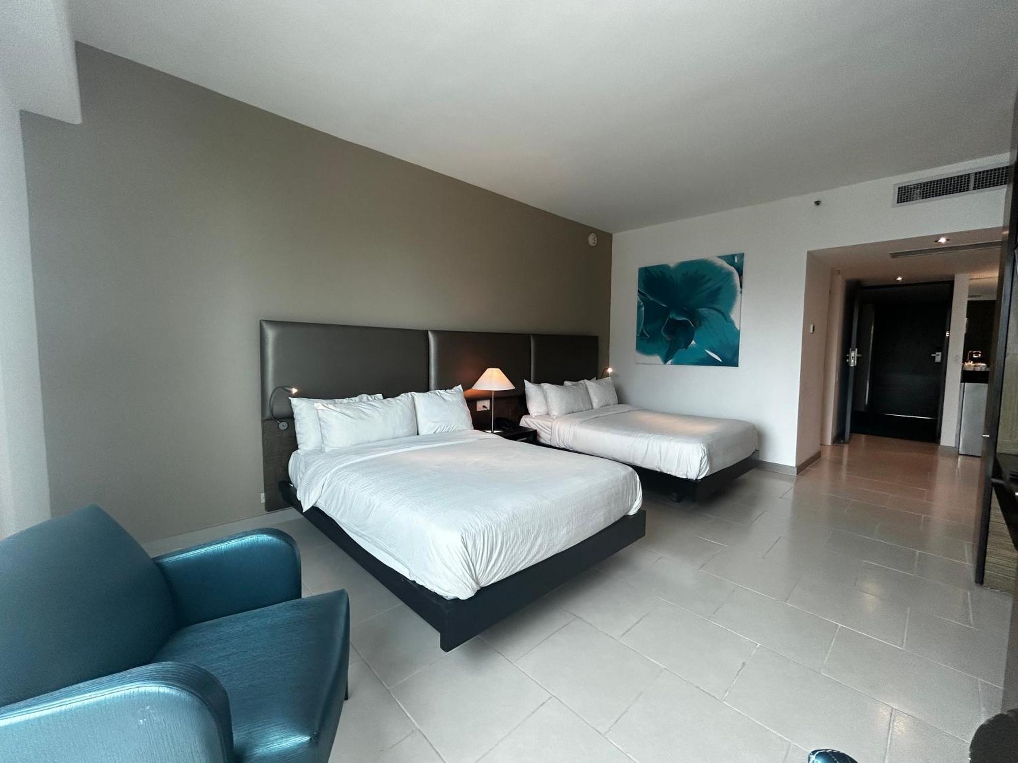 Decapolis Hotel ปานามาซิตี้ ภายนอก รูปภาพ The photo shows a spacious hotel room featuring two queen-sized beds, each with white bedding. There is a blue armchair in one corner, adding a pop of color to the neutral-toned decor. A large piece of artwork with a blue flower is displayed on the w