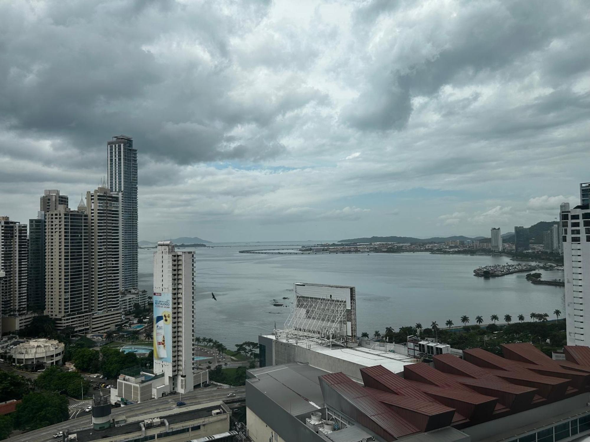 Decapolis Hotel ปานามาซิตี้ ภายนอก รูปภาพ The photo shows a coastal cityscape featuring tall buildings and skyscrapers lining the waterfront. In the foreground, there are modern structures with distinctive architectural designs. The scene is dominated by a cloudy sky, suggesting an overcast 