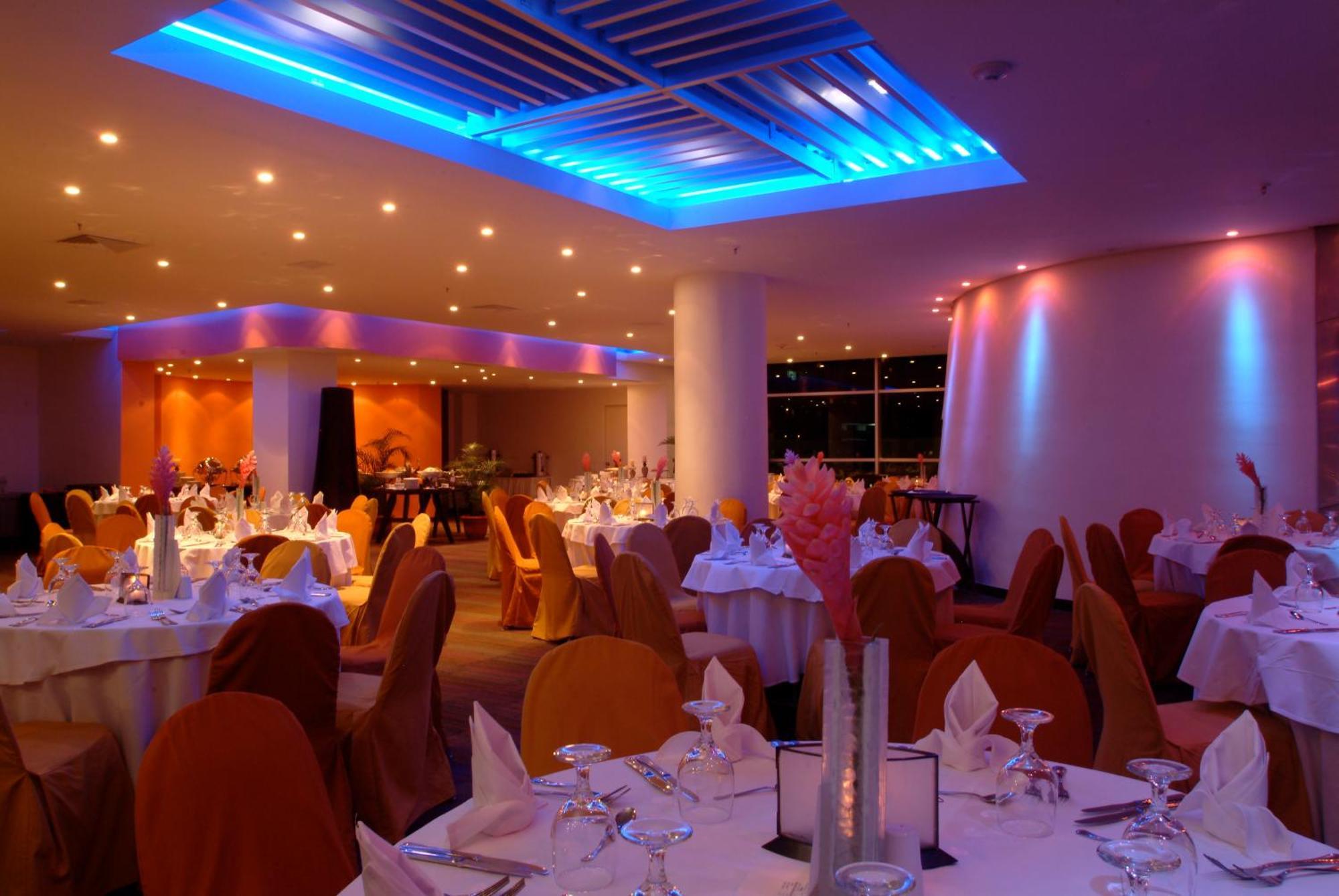 Decapolis Hotel ปานามาซิตี้ สิ่งอำนวยความสะดวก รูปภาพ The image shows an elegant restaurant interior. The dining area features round tables covered with white tablecloths, and each table is set with glassware and napkins. The chairs are draped in orange fabric, contributing to a warm and inviting atmosp