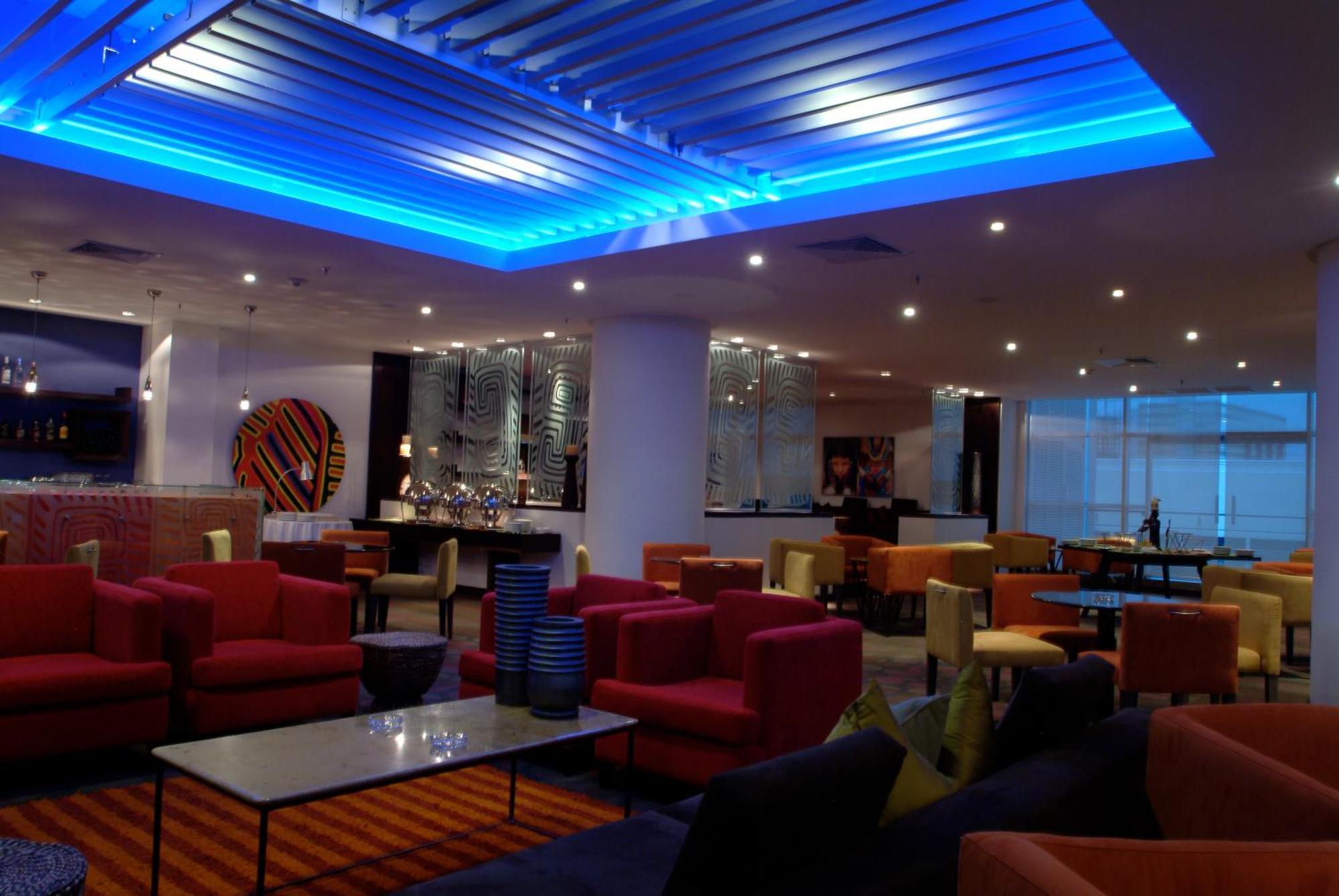 Decapolis Hotel ปานามาซิตี้ ร้านอาหาร รูปภาพ The photo shows a modern lounge or lobby area with various seating arrangements. The furniture includes colorful chairs and sofas, predominantly in shades of red, pink, and yellow, arranged around glass-topped tables. The ceiling features a blue illu